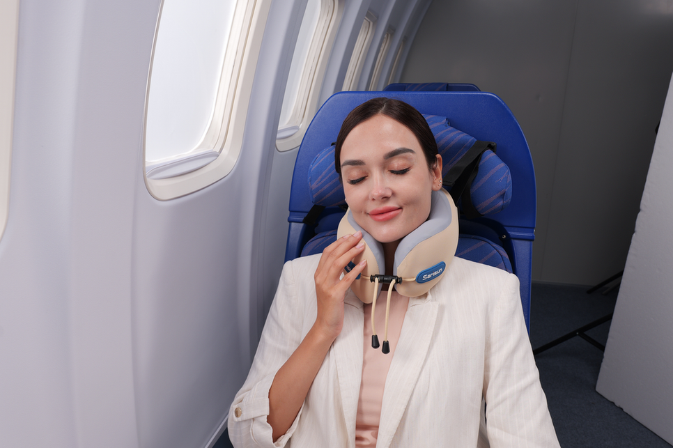 A woman resting with her eyes closed in an airplane seat while gently touching her beige travel pillow.