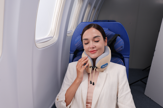 A woman resting with her eyes closed in an airplane seat while gently touching her beige travel pillow.