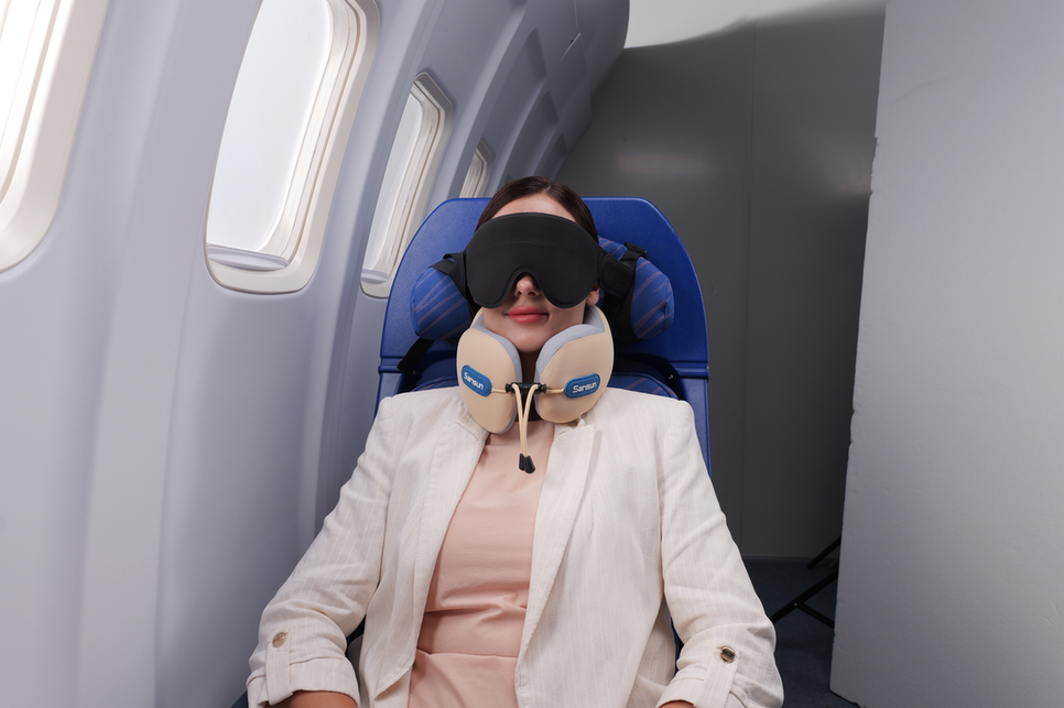 A woman resting comfortably in an airplane seat while wearing a black sleep mask and a securely fastened travel pillow around her neck.