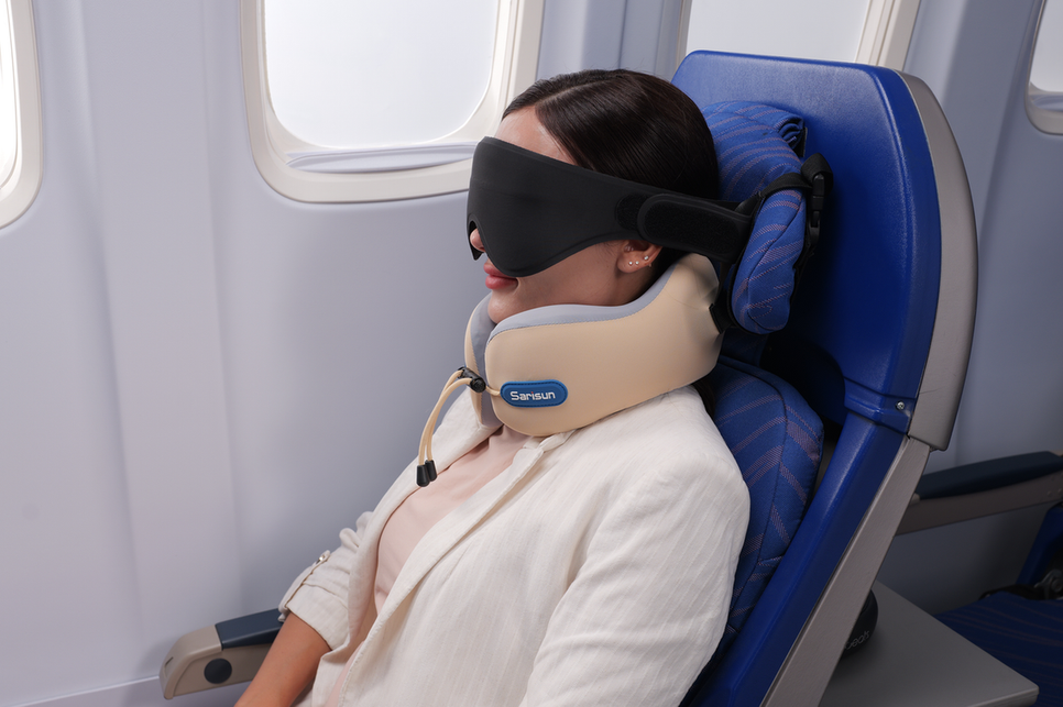 Passenger resting by an airplane window while wearing an eye mask and travel neck pillow